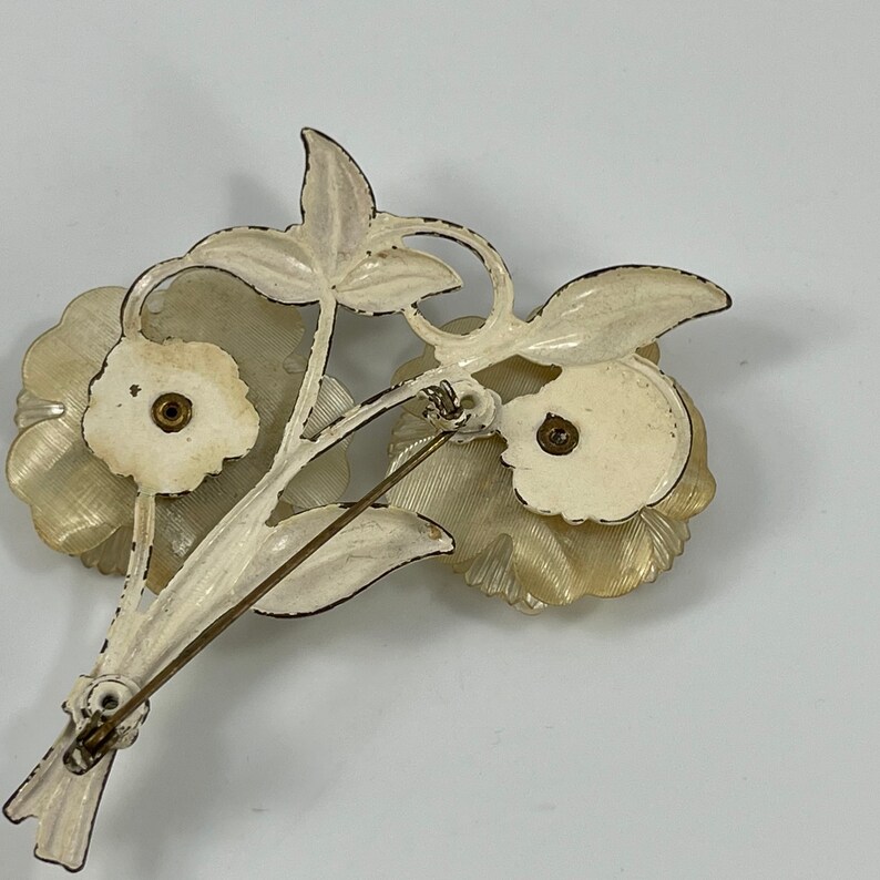May include: White metal flower brooch with two white flowers and a gold pin. The back of the brooch is visible.