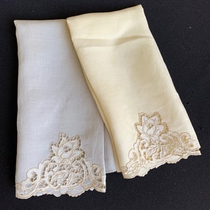 May include: Two embroidered linen tea towels, one white and one light beige, with a floral design on the bottom edge.