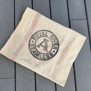 May include: A vintage, off-white canvas bag with red and blue stripes. The bag has a black circular stamp that reads "Royal River Seamless" with a large letter "A" in the center.