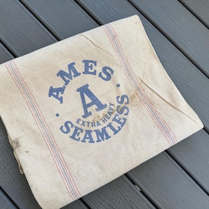 May include: Cream-colored fabric with blue text that reads "AMES A EXTRA HEAVY SEAMLESS". The fabric has red and blue stripes running vertically. The fabric appears to be vintage or antique.