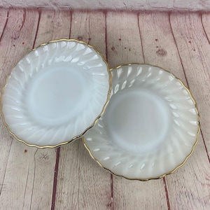 May include: Two white glass plates with a swirl design and gold trim. The plates are round with a scalloped edge. The plates are on a light brown wooden surface.