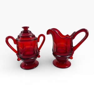 Puede incluir: Un azucarero de cristal rojo con tapa y una jarra de crema a juego. Ambas piezas tienen asas curvas y detalles decorativos. El azucarero tiene una tapa con un pequeño pomo. La jarra de crema tiene un pico vertedor.