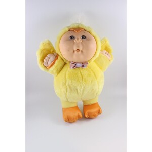 Pode incluir: Uma boneca de pelúcia vintage com uma fantasia de pato amarelo. A boneca tem um rosto redondo com olhos grandes, um nariz pequeno e uma expressão de surpresa. Usa uma gravata borboleta rosa e detalhes laranja nos pés e nas mãos. A boneca está em um fundo branco.
