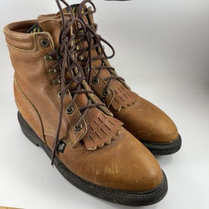 May include: A pair of brown leather work boots with laces and fringe detailing. The boots have a worn appearance, with visible scuffs and a black sole. The boots are designed for work and outdoor activities.
