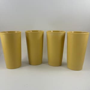 May include: Four tall, yellow drinking glasses. The glasses are made of a solid, opaque material and have a slightly textured surface. They are cylindrical in shape with a slight taper towards the base. The glasses are empty and arranged in a row.