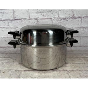 Puede incluir: Olla de cocina de acero inoxidable con tapa. La olla tiene dos asas negras a cada lado. La tapa también tiene un asa negra a cada lado. La olla está sobre un fondo de ladrillos blancos y grises.