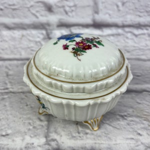 May include: A white porcelain trinket box with a floral design on the lid. The box has three gold-painted feet and a gold trim around the top edge.