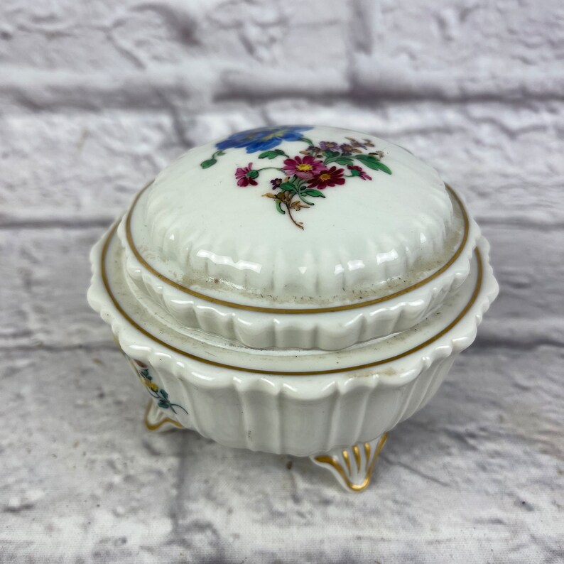 May include: A white porcelain trinket box with a floral design on the lid. The box has three gold-painted feet and a gold trim around the top edge.