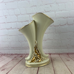Puede incluir: Un jarrón de cerámica blanca con detalles dorados. El jarrón tiene dos aberturas en forma de trompeta y una base decorativa.