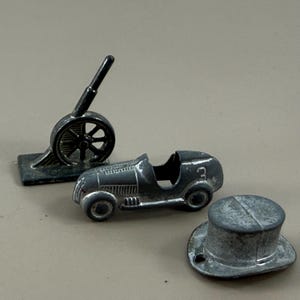 May include: Three small, antique metal toys: a cannon, a race car with the number 3, and a top hat. The toys are silver-colored and show signs of wear, suggesting they are vintage items. The background is a neutral, light brown.