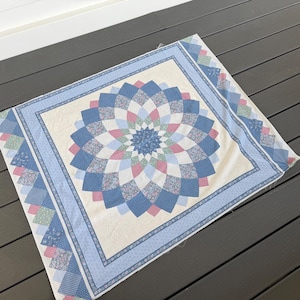May include: A floral patterned quilt with a central design resembling a flower. The quilt features a cream-colored center with a blue, pink, and white floral pattern. A light blue border surrounds the central design, with a patterned edge.