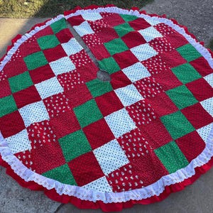 May include: A circular Christmas tree skirt featuring a patchwork design of red, green, and white fabric squares. The skirt has a central opening, a white eyelet lace trim along the outer edge, and a red ruffled border surrounding the lace.