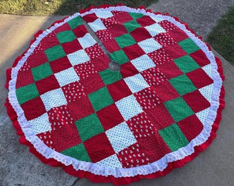 May include: A circular Christmas tree skirt featuring a patchwork design of red, green, and white fabric squares. The skirt has a central opening, a white eyelet lace trim along the outer edge, and a red ruffled border surrounding the lace.