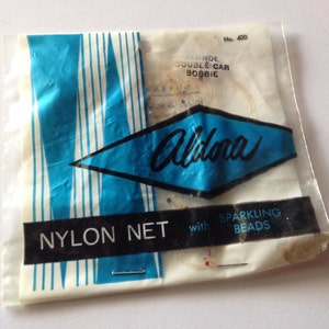 May include: A white plastic bag with blue and white stripes. The bag contains a package of blonde double-cab bobbie hair with sparkling beads. The bag has the text "Aldona Nylon Net with Sparkling Beads" printed on it.