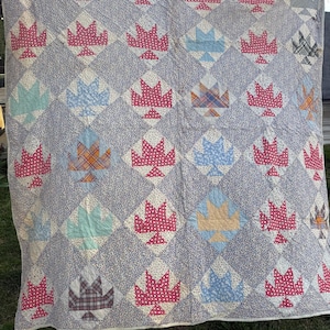May include: A large, handmade quilt with a repeating pattern of red maple leaf shapes on a light blue background. The quilt features various colors of maple leaves, including red, blue, and plaid patterns. The quilt is hanging outside on a clothesline.