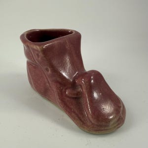 May include: A burgundy-colored ceramic boot-shaped vase. The vase has a rounded toe and a flared opening at the top. The boot has a textured surface and a slightly glossy finish. The vase is likely a decorative item.