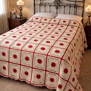 May include: Cream-colored crochet blanket with a red square pattern and red circular accents. The blanket is draped over a bed with a decorative metal headboard. The room has a vintage aesthetic.