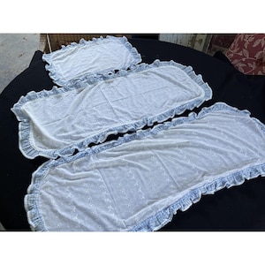 May include: Three white eyelet lace pieces with blue trim and ruffled edges. The pieces are rectangular and appear to be decorative linens, possibly for a bed or table. The eyelet pattern is visible throughout the fabric.