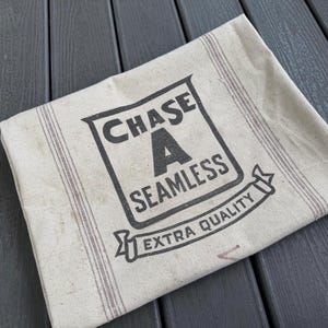 May include: A folded, off-white fabric with a printed logo that reads "CHASE A SEAMLESS EXTRA QUALITY" in black. The fabric has vertical stripes in a muted red-brown color. The background is a dark gray wooden surface.