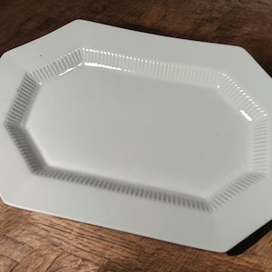 May include: A white, octagonal serving platter with a ribbed border. The platter has a smooth, glossy surface and is set on a wooden surface. The design is simple and elegant, suitable for various culinary presentations.