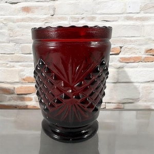 Peut inclure: Un vase en verre rouge foncé avec un motif en diamant. Le vase a un bord festonné et une base large.