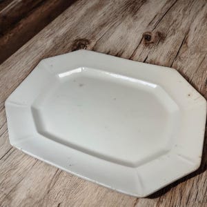 May include: A white, octagonal serving platter with a slightly raised rim. The plate has a glossy finish and is set against a rustic wooden surface. The platter is a classic piece for serving food.
