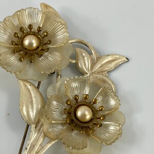 May include: A vintage brooch featuring two white flowers with pearl centres and gold accents. The flowers are set on a white metal base with a leaf design.