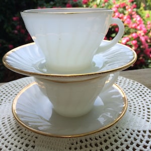 May include: Two white teacups and saucers stacked on top of each other. Each cup and saucer has a gold trim. The cups have a ribbed design. The background is a blurred view of a garden with pink flowers.