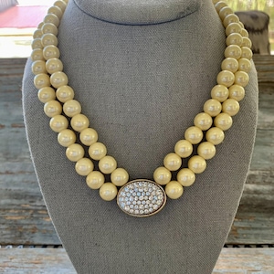 Puede incluir: Collar de doble hilera en crema con cuentas redondas grandes. El collar tiene un colgante ovalado dorado con pequeños cristales brillantes. Se muestra sobre un maniquí gris.