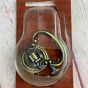 May include: A gold-toned metal cabinet knob with a decorative ring and a hook. The knob is packaged in a clear plastic blister pack on a brown card with the text "Trim Furniture Trim".