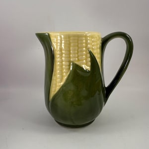 May include: A green and yellow ceramic jug shaped like an ear of corn. The jug has a handle and a spout.