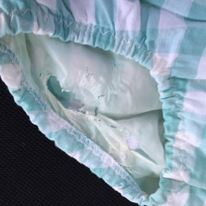 May include: A close-up of a green and white gingham fabric with a hole in the fabric. The fabric is gathered at the top and has a white seam.