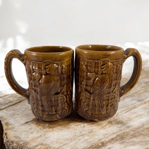 Pode incluir: Duas canecas de cerâmica marrom com um design em relevo de pássaros e folhas. As canecas têm uma alça no lado.