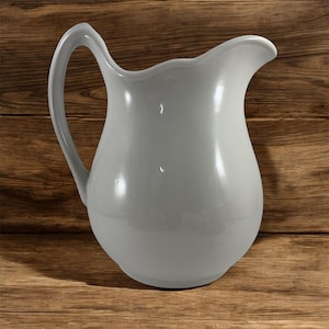 May include: A gray ceramic pitcher with a curved handle and a scalloped rim. The pitcher has a glossy finish and is set against a wooden background. This decorative pitcher is suitable for serving beverages.