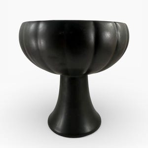 Può includere: Una ciotola nera su piedistallo con bordo scanalato. La ciotola poggia su una base svasata, creando una silhouette classica ed elegante. Il colore scuro e il design semplice lo rendono un pezzo versatile per l'arredamento della casa.
