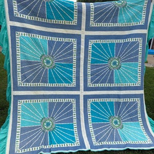 Op de afbeelding: Een grote, handgemaakte quilt met een geometrisch ontwerp in blauw- en turkooistinten. De quilt heeft een centraal sunburst-patroon in elke vierkant, met een groen en wit bloemencentrum. De quilt wordt buiten tentoongesteld, met een gefranjerde rand.