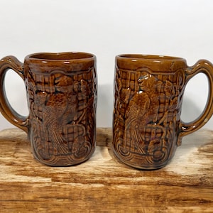 Pode incluir: Duas canecas de cerâmica marrom com um design em relevo de um pássaro e um padrão xadrez. As canecas têm uma alça do lado direito.