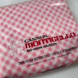 May include: A pink and white gingham twin fitted bottom sheet in its original packaging. The packaging features the brand name "Cannon Monticello" and the text "No-Iron Muslin". The sheet is designed for a 39 x 76 inch mattress.