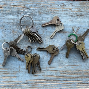 Op de afbeelding: Een verzameling vintage sleutels, sommige met sleutelringen, in verschillende maten en afwerkingen, waaronder zilver, messing en koper. De sleutels zijn gerangschikt op een lichtblauw houten oppervlak.