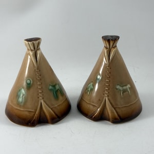 May include: Two ceramic salt and pepper shakers in the shape of teepees. The shakers are tan with brown detailing and feature green and brown decorative elements. The design includes a dog and other shapes.