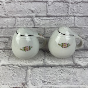 Może przedstawiać: Dwie białe solniczki i pieprzniczki ceramiczne. Każda ma uchwyt i srebrną obwódkę. Ozdobione są kwiatowym wzorem z różowych i zielonych kwiatów. Idealne na stół.