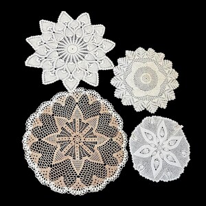 May include: Four white and beige crochet doilies of varying sizes and designs are displayed against a black background. The doilies feature intricate patterns, including floral and geometric shapes, showcasing the delicate craftsmanship.