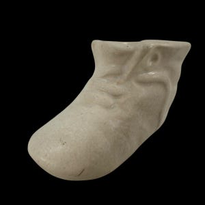 Pode incluir: Pequena figura de bota de cerâmica esbranquiçada. A bota tem uma ponta arredondada e uma textura ligeiramente enrugada, com um acabamento liso. A bota está sobre um fundo preto.