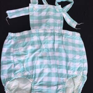 May include: A light blue and white gingham baby onesie with straps. The onesie has a ruffled bottom and is made of a soft, comfortable fabric.