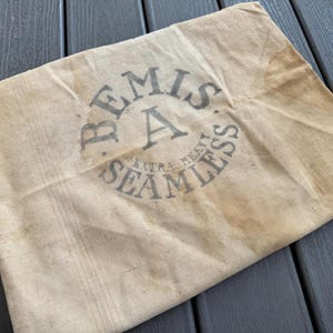 Puede incluir: Una bolsa beige Bemis Extra Heavy Seamless. La bolsa tiene un aspecto vintage descolorido con un logotipo circular en el centro. El logotipo presenta el nombre "Bemis" y las palabras "Extra Heavy Seamless".