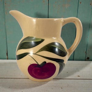 Puede incluir: Jarra de cerámica color crema con un asa grande y un pico. La jarra está decorada con una cereza roja oscura y hojas verdes y negras. La jarra probablemente se utiliza para servir bebidas.