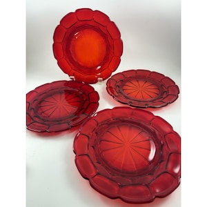 Puede incluir: Cuatro platos de vidrio rojo con borde festoneado y diseño de rayos de sol en el centro. Los platos son de un tono rojo vibrante y parecen ser de vidrio. Los platos están dispuestos sobre una superficie blanca.