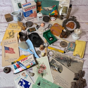 May include: A selection of vintage collectables, including a Band-Aid tin, postcards, a clown figurine, and a box of Crayola crayons. Other items include a small jug, a comb, and various trinkets, all arranged on a light-coloured surface.