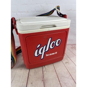 May include: A red Igloo Tag-Along 10 cooler with a white lid and a rainbow-striped shoulder strap. The cooler has the Igloo logo in white script. The cooler is designed to keep items cold.