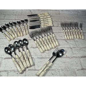 Pode incluir: Uma coleção de talheres vintage, incluindo colheres, garfos e facas. Os talheres apresentam cabos creme com um design floral decorativo. Os utensílios de aço inoxidável estão dispostos sobre uma superfície de tijolo cinza claro.